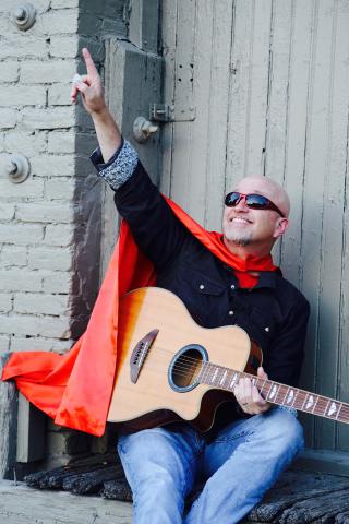 Roger Day in a cape holding a guitar and pointing to the sky